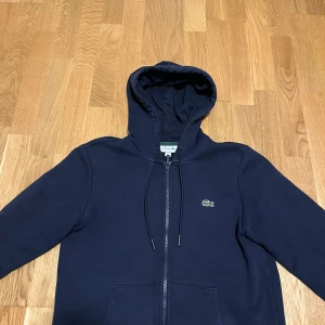 Marinblå hoodie från Lacoste - Snygg marinblå hoodie från Lacoste med dragkedja och klassisk krokodillogga på bröstet. Tröjan har huva med snörning, långa ärmar och två fickor framtill. Perfekt för dig som gillar stilren och sportig look. Ny pris 1800kr köpte den på Åhléns 
