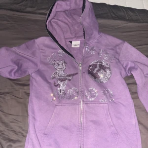 Lila hoodie Aim For The Moon med glitter - Lila hoodie med dragkedja från Aim For The Moon. Framsidan har glittriga detaljer, en tecknad figur och text samt motiv av jordklot och peace-symbol. Utrustad med huva och två fickor framtill. Perfekt för dig som gillar statement-plagg med extra bling.