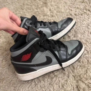 Nike Air Jordan 1 Mid Svart/Grå - Snygga Nike Air Jordan 1 Mid sneakers i svart och grått skinn med röda detaljer på sidan och tungan. Klassisk siluett med svart swoosh, perforerad tå och svart snörning. Perfekta för dig som vill ha en ikonisk och stilren sneaker med streetkänsla.