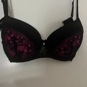 Svart och rosa spets-bh med smycke - Säljer en snygg bh i svart med rosa underton och detaljerad spets över kuporna. Bh:n har justerbara axelband, liten rosett och ett glittrande smycke framtill. Sidorna är också täckta av spets för extra stilpoäng. SKORLEK 85D