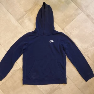 Mörkblå hoodie från Nike - Hej! Jag säljer en mörkblå hoodie från Nike med klassisk vit logga på bröstet. Tröjan har luva, långa ärmar och en stor magficka framtill. Materialet är mjukt och skönt. Har dessutom bomull inuti för en ännu skönare känsla. Den är i storlek 147-158. De medföljande mjukisbyxorna finns också att köpa. Perfekt för dig som gillar sportig och avslappnad stil! Tveka inte att höra av dig vid frågor!