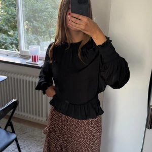 Svart blus med volang och puffärm - Supersnygg svart blus med volangdetalj nertill och puffiga långärmar. Blusen har rund halsringning och kan knytas i nacken. Kan stylas både med volang och utan till jeans eller kjol. Perfekt på kontoret! 