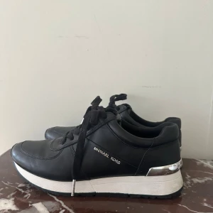 Svarta sneakers från Michael Kors - Stilrena svarta sneakers från Michael Kors i skinn med silver sulor och silverdetaljer vid hälen. Skorna har snörning och diskret logga på sidan. Perfekta för dig som gillar en clean och modern look med exklusiv känsla.
