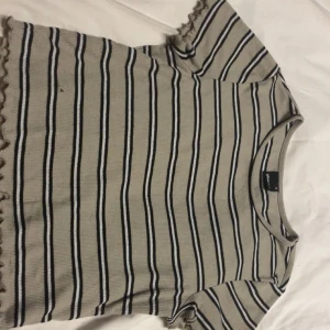 Randig ribbad topp från Gina Tricot - Säljer en beige och svart ribbad topp från Gina Tricot med smala svarta och vita ränder. Toppen har korta ärmar och vågiga kanter vid ärmslut och nederkant. Perfekt för dig som gillar en enkel men ändå trendig stil.