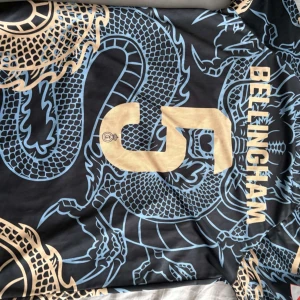 Real Madrid Bellingham draktröja Adidas - Snygg Real Madrid fotbollströja med Bellingham 5 på ryggen. Svart bas med guldiga och ljusblåa drakdetaljer, klubbmärke och Adidas-logga framtill. Kortärmad modell i lätt och ventilerande material, perfekt för match eller träning. PRIS KAN DISKUTERAS 