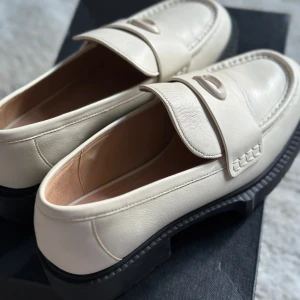 Coach Leah Loafer i ljusbeige skinn - Snygga ljusbeiga loafers från Coach i modellen Leah Loafer. Skorna är tillverkade i slätt skinn med chunky svart sula och dekorativ detalj på ovandelen. Perfekta för dig som vill ha en stilren men trendig look. Kommer i originalkartong.
