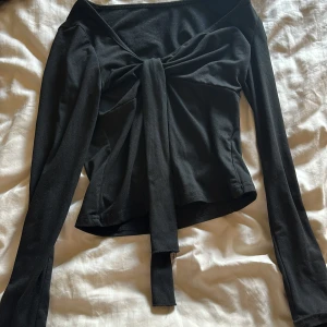 Svart långärmad topp med knyt - Säljer en svart långärmad topp med snygg knyt-detalj framtill. Toppen har en croppad passform och är tillverkad i mjukt bomullsliknande material. Perfekt för dig som gillar trendiga och stilrena plagg med det lilla extra.