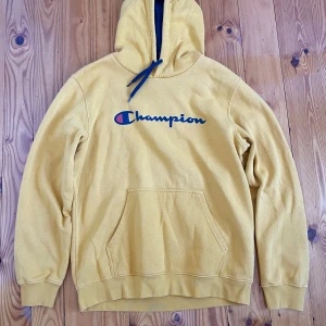 Champion hoodie gul - Säljer en gul hoodie från Champion med stor logga på bröstet. Tröjan har huva med blå snörning och en klassisk magficka. Perfekt för dig som gillar streetwear och vill ha en färgglad look.