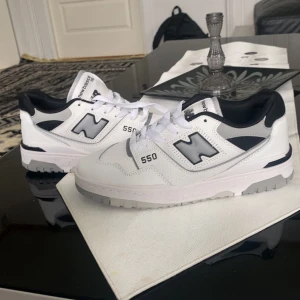 New Balance 550 vita sneakers - Snygga New Balance 550 sneakers i vitt med svarta och grå detaljer. Klassisk siluett med rund tå, snörning och platt sula. Ovandel i skinn och syntet, med tydlig New Balance-logga på sidan. Perfekta för dig som gillar stilrena och sportiga skor.