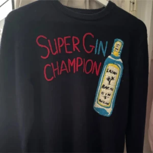 Svart stickad tröja Super Gin Champion - Svart stickad tröja från Saint Barth med broderad text 'Super Gin Champion' i rött och blått samt en stor broderad ginflaska på framsidan. Tröjan har rund hals och långa ärmar, perfekt för dig som gillar unika statement-plagg.