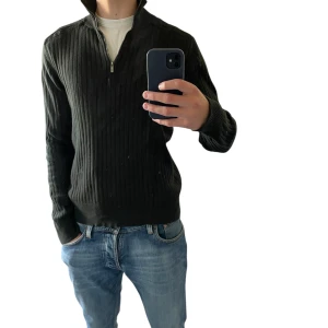 Svart ribbad tröja från Calvin Klein | M - Svart ribbstickad tröja från Calvin Klein Jeans med halv dragkedja och hög krage. Tröjan har lång ärm och en clean, stilren look som passar perfekt till jeans. Materialet är mjukt och skönt, perfekt för kyligare dagar. Tröjan har en liten fläck nära dragkedjan som knappt märks