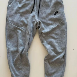 Grå mjukisbyxor från Nike - Snygga grå mjukisbyxor från Nike med svart logga på vänster ben. Byxorna har dragsko i midjan och är tillverkade i mjukt bomullsmaterial. Perfekta för chill eller träning, med en skön och avslappnad passform.