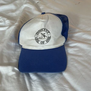 Blå och vit keps Innocent One of One - Snygg truckerkeps i blått och vitt från Innocent One of One. Framsidan har ett tryckt motiv med text och ängel, medan baksidan är i blå mesh för extra ventilation. Perfekt för dig som vill sticka ut med en unik och cool keps.