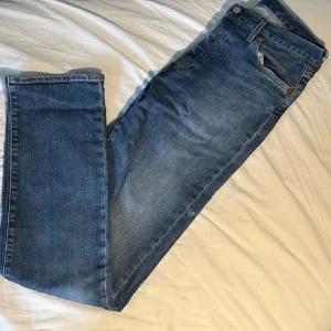 Levi's 501 blå jeans 30/34 (män) - Klassiska Levi's 501 jeans i mellanblå tvätt med raka ben och normal passform. Jeansen har fem fickor, knappgylf fast med ett litet hål på sidan som man lätt kan sy ihop om man är lagd åt de hållet. Perfekta för dig som gillar tidlös denimstil.