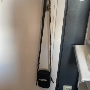 Svart No Fear axelväska med logga - Svart axelväska från No Fear med vit broderad logga framtill. Väskan har justerbar axelrem med No Fear-tryck, två dragkedjefack och detaljer i silverfärgad metall. Perfekt storlek för mobil och småprylar, sportig och streetig vibe.