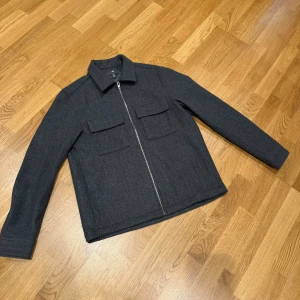 Grå overshirt/Jacka - Stilren grå overshirt från H&M i ullmix med klassisk krage, två stora bröstfickor med lock och hel dragkedja framtill. Jackan har långa ärmar med knapp vid ärmslut och en rak, enkel passform. Perfekt för lager-på-lager under kyliga dagar. Andvänd max 5 gånger, säljer pga för liten. Nytt pris 499! Pris kan diskuteras vid snabb affär☺️