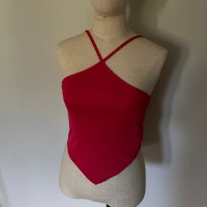 Röd asymmetrisk croppad linne-topp - Säljer en röd croppad linne-topp med asymmetrisk skärning och smala axelband som korsas framtill. Toppen har en spetsig nederkant och är figurnära, perfekt för dig som gillar trendiga och unika plagg. Materialet känns mjukt och stretchigt.