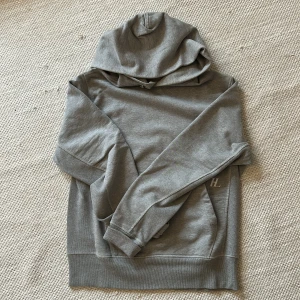 Grå hoodie från Helmut Lang med HL-logga - Säljer en stilren grå hoodie från Helmut Lang i 100% bomull. Tröjan har stor vit HL-logga på ryggen, huva och klassisk känguruficka framtill. Perfekt för dig som gillar minimalistisk design och vill ha något bekvämt och snyggt.