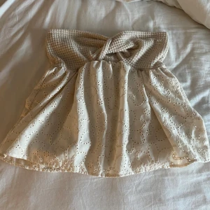 Beige broderad tubtopp med twist - Supersöt beige tubtopp med twistad smock upptill och broderade blommor på hela tyget. Toppen är ärmlös och har en luftig, flowy passform. Perfekt för varma dagar och ger en romantisk vibe till din outfit.