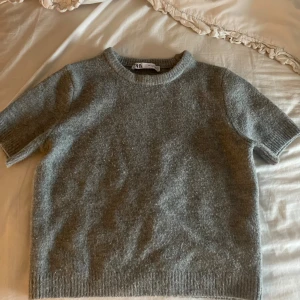 Grå stickad tröja från Zara - Mysig grå stickad tröja från Zara med korta ärmar och rund halsringning. Tröjan har en enkel och stilren design med ribbade kanter vid hals, ärmslut och nederkant. Perfekt för lager-på-lager och chill dagar.