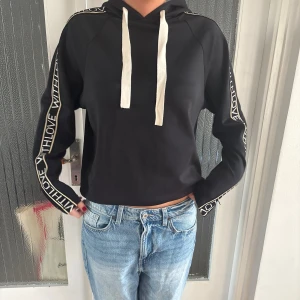 Svart hoodie med textdetaljer från maje - Svart hoodie med vita snören och texten 'WITH LOVE' längs båda ärmarna. Tröjan har huva och långa ärmar, samt en sportig look med kontrasterande detaljer. Perfekt för en chill och avslappnad stil. Från maje och super härligt material. Har inte kvar kvittot, finns matchande byxor. Skulle säga att de är en S. 