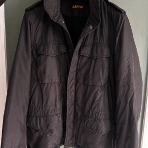 Svart vindjacka från Aspesi, M - Svart vindjacka från Aspesi i storlek M. Jackan har axelklaffar och dragkedja samt knappar. Jackan är gjord i ett lätt, syntetiskt material som ger en clean och stilren look. Små defekter på en knapp och intill i jackan (se bilderna)