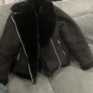 Svart pälsjacka med dragkedjor - Säljer en svart pälsjacka med bred krage och detaljer i fuskpäls. Jackan har silvriga dragkedjor framtill och på fickorna samt spänndetaljer vid midjan. Perfekt för kalla dagar och ger en riktigt cool vibe.