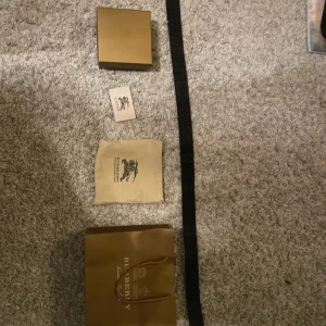 Burberry bälte med klassiskt rutmönster - Säljer ett snyggt Burberry bälte med det ikoniska rutmönstret i beige, rött och svart. Bältet har ett stort metallspänne med Burberrys TB-logga i svart. Kommer med originalpåse, box och Burberry-påse. Perfekt accessoar för att lyfta din outfit.