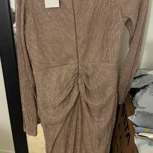 Champagnefärgad glittrig långklänning - Supersnygg långklänning från Maniju  i champagnefärgat glittrigt tyg. Klänningen har v-ringning, långa ärmar och omlott-detalj framtill med snygg drapering. Perfekt för dig som vill sticka ut med en elegant och trendig look. Helt ny och oanvänd. Köpte den för 1500 säljer den för 600