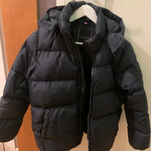 Mörkblå Essential down jacket från Tommy Hilfiger - Mörkblå  Essential down jacka från Tommy Hilfiger med huva och dragkedja. Jackan har en klassisk  look och är vadderad för extra värme. Insidan har logotypen broderad i nacken och diskreta röda och vita detaljer längs fodret. Perfekt för kalla dagar. Jackan har en liten reva i ryggen.