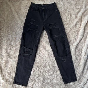Svarta mom jeans från PrettyLittleThing - Svarta mom jeans från PrettyLittleThing med stora slitningar och råa kanter på benen. Jeansen har hög midja och klassisk femficksmodell. Innerbenslängd 76 cm, midja tvärsöver 36 cm