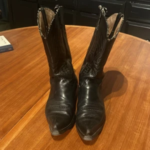 Svarta cowboy boots i skinn, storlek 42 - Säljer ett par svarta cowboy boots i äkta skinn med klassisk spetsig tå och låg klack. Bootsens skaft har snygga broderade detaljer och insidan är i naturfärgat läder. Perfekt för dig som vill ha en cool westernstil och gillar robusta boots.