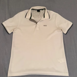 Boss Piké  - Snygg vit pikétröja från BOSS med klassisk krage och diskreta svarta och gröna detaljer på krage och ärmslut. Tröjan har korta ärmar och broderad BOSS-logga på bröstet. Perfekt för en stilren och fräsch look.