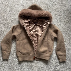 Alessa furjacket satin beige  - Mysig beige stickad jacka från Alessa med huva kantad av mjuk fuskpäls. Jackan har ribbade muddar, två fickor framtill och dragkedja med läderdetalj. Insidan är fodrad i glansigt tyg för extra komfort. Perfekt för kyliga dagar.