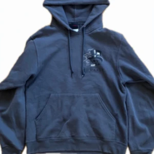 Mörkgrå Ed Hardy hoodie med tryck - Säljer en mörkgrå hoodie från Ed Hardy med stor grafisk döskalle och hatt-tryck på ryggen samt mindre tryck på bröstet. Tröjan har huva med snörning och en klassisk magficka. Perfekt för dig som gillar en lite mer alternativ stil. Hör gärna av er vid frågor!