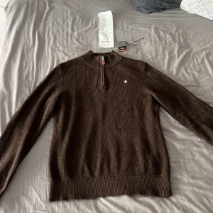 Brun stickad tröja Morris med half zip - Säljer en brun stickad tröja från Morris med ribbad struktur och half zip-dragkedja med läderdetalj. Tröjan har lång ärm, hög krage och en broderad logga på bröstet. Perfekt för lager-på-lager under hösten och vintern.
