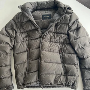 Pufferjacka från Massimo Dutti - Snygg olive/grå pufferjacka från Massimo Dutti med huva och två dragkedjeförsedda fickor. Jackan har quiltad design och stängs med dragkedja. Perfekt för kyliga dagar och ger en clean, modern look.