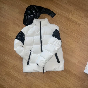 Vit och svart pufferjacka Moose Knuckles - Säljer en fet pufferjacka från Moose Knuckles i vitt med svarta detaljer och avtagbar huva. Jackan har glansiga svarta partier på ärmar och rygg, dragkedja framtill och två fickor med svarta dragkedjor. Perfekt för kalla dagar när du vill sticka ut. Storlek M och den är aldrig använd