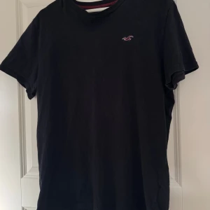 Mörk marinblå t-shirt från Hollister, storlek L - Mörk marinblå t-shirt från Hollister med klassisk crew neck och diskret röd logga på bröstet. Modellen är enkel och stilren, perfekt för dig som gillar en clean look. Tillverkad i mjuk bomull som känns skön mot huden. Passar dig som vill ha en basic tee med lite extra vibe. T-shirten är lite nopprig, men man ser inte det lika tydligt i verkligheten