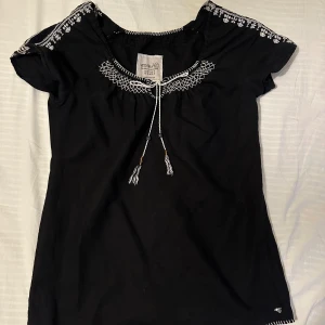 Svart blus med broderi från Esprit - Svart blus från Esprit med vita broderade detaljer längs halsringning och axlar. Blusen har korta ärmar, knytband med tofsar framtill och ett litet broderat Esprit-logo nertill. Tillverkad i mjuk bomull och linne och har en avslappnad passform.