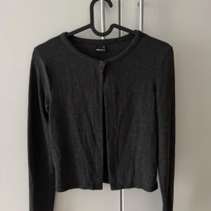 Mörkgrå kofta från Gina Tricot - En enkel och stilren mörkgrå kofta från Gina Tricot i storlek XS. Koftan har rund halsringning, lång ärm och en knapp framtill. Perfekt att slänga över en topp när det blir lite kyligt. Tunn och mjuk i materialet. Koftan är hel ny och har aldrig används.