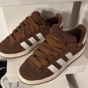 Bruna Adidas Campus sneakers mocka - Snygga bruna Adidas Campus sneakers i mocka med klassiska vita ränder på sidorna och breda bruna skosnören. Skorna har en platt sula och rund tå, perfekt för dig som gillar retrostil och streetwear. Adidas logga på plösen ger extra vibe.