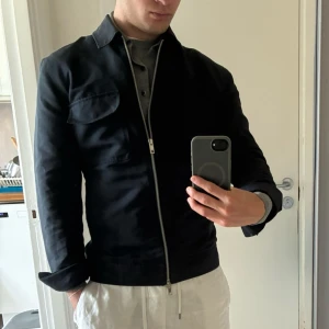 Mörkblå overshirt från Elvine, storlek S - Snygg svart overshirt från Elvine i stilren design. Jackan har klassisk krage, två bröstfickor med lock och hel dragkedja framtill. Tillverkad i bomullsmix som ger en clean och modern look. Perfekt för lager-på-lager och passar till det mesta. Nyskick!