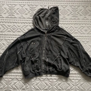 Thug Club hoodie jacket - Cool svart hoodie med råa sömmar, hel dragkedja och huva med nitar. Framsidan har två silvriga metalldetaljer, en stjärna och en måne. Hoodien är croppad och har en loose passform, perfekt för en edgy streetstyle-look. Fullzip till nacken