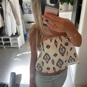 Vit och svart mönstrad linnetopp - Säljer en luftig vit linnetopp med svart grafiskt mönster. Toppen har smakt axelband som knyts på ena axeln och en croppad passform. Perfekt för varma dagar och ger en chill vibe till din outfit.