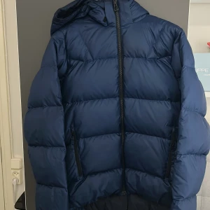 Blå pufferjacka från Helly Hansen - Säljer en marinblå pufferjacka från Helly Hansen med huva och hel dragkedja framtill. Jackan har två snedställda fickor med dragkedja och är vadderad för extra värme. Perfekt för kalla dagar och snygg till streetwear. Den är använd 2 vintar sitter större än vad man tror.