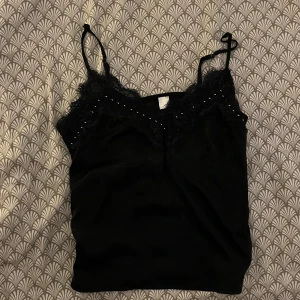 Svart linnetopp med spets och pärlor - Snygg svart linnetopp med tunna axelband och dekorativ spets längs urringningen. Små silvriga pärlor pryder spetsen för extra detalj. 