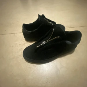 Svarta Arigatos,mocka zise 39 - Arigatos i mocka med coola dragkedjor framtill istället för klassisk snörning. Skorna har låg profil och platt sula, vilket ger en clean och modern look. Perfekta för dig som gillar stilrena detaljer och vill sticka ut lite extra.