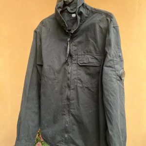 Svart overshirt från C.P. Company - Svart overshirt från C.P. Company med dragkedja framtill, bröstficka med lock och den klassiska linsen på ärmen. Jackan har hög krage och är tillverkad i bomull. Perfekt för lager-på-lager och en clean streetwear-look. Storklek L men sitter som S.