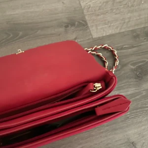 Röd quiltad axelväska från Valentino - Säljer en snygg röd axelväska från Valentino med quiltad design och stort logotyptryck framtill. Väskan har en kedjerem med röda detaljer och guldiga inslag. Materialet är mjuk sammet och insidan har flera fack och dragkedja. Perfekt för dig som vill sticka ut med en trendig accessoar.